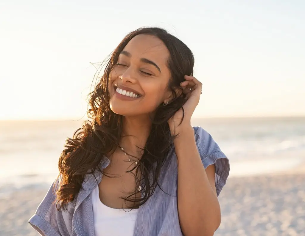 Smiling latin hispanic woman relaxing on beach with closed eyes at sunset. Beautiful mixed race woman enjoying wind fluttering hair. Charming and calm young woman breathing fresh air at summer beach with copy space.