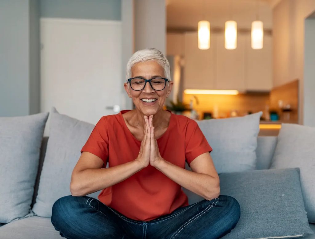 Attractive mature woman meditating at home. Calm senior woman relax on couch meditating in lotus position, peaceful aged female practice yoga at home relieve stress or renew energy, elderly lady do physical exercises following healthy lifestyle