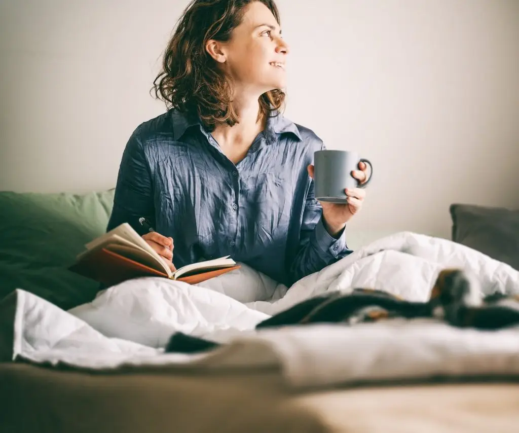 Young cheerful woman sitting on bed writing in diary and drinking morning coffee