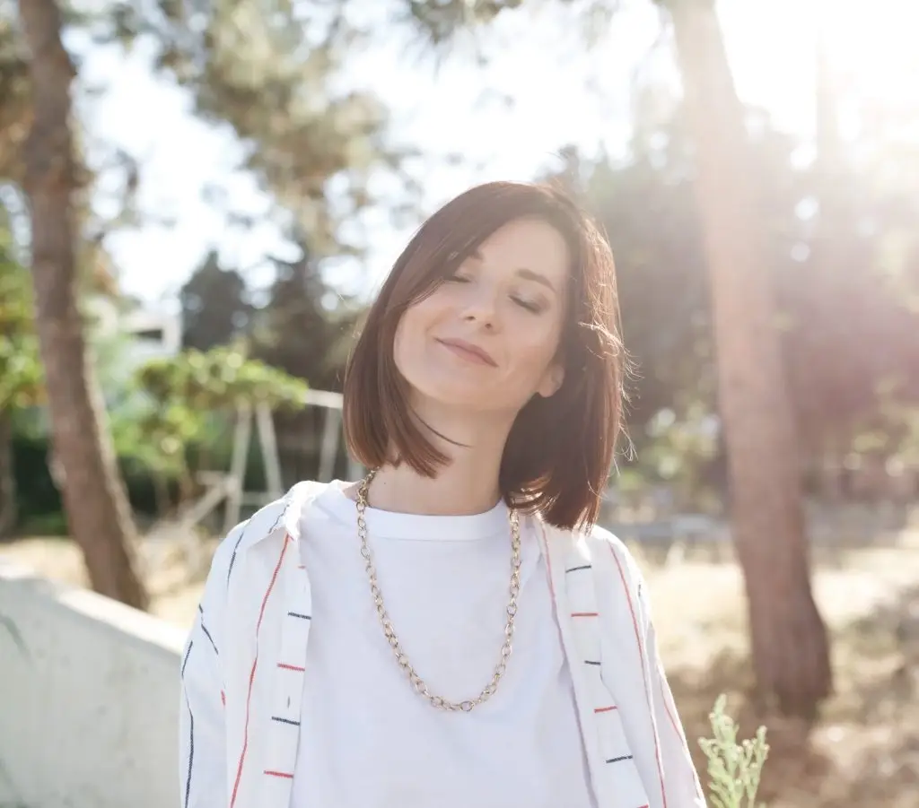 Close up outdoors portrait of a young and beautiful woman in white clothes, smiling with her eyes closed, sunlight touch her neck, blurred nature background.Freedom concept