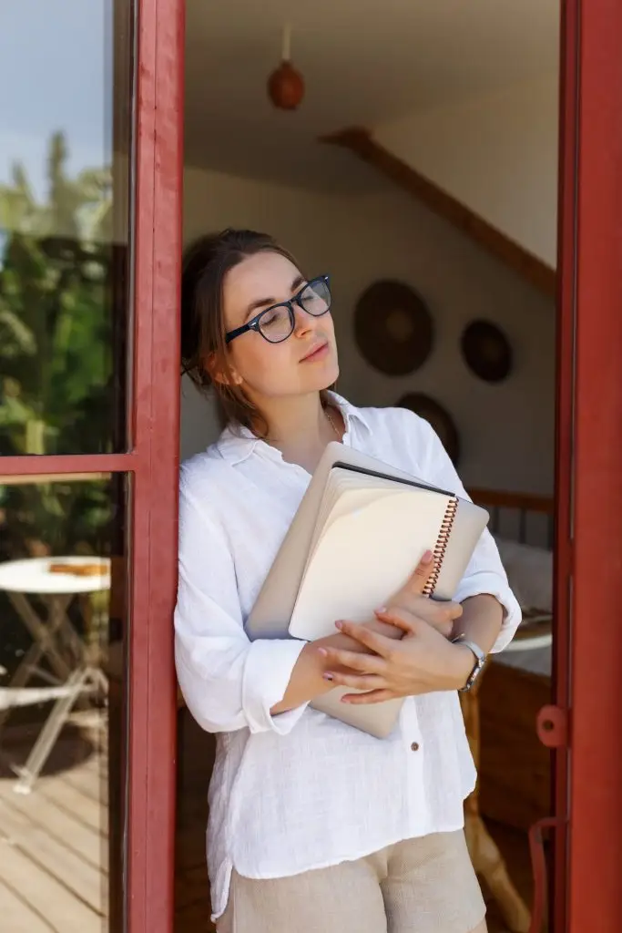 Portrait of businesswoman or student with folder and laptop in hand standing near the glass door background and looking forward.