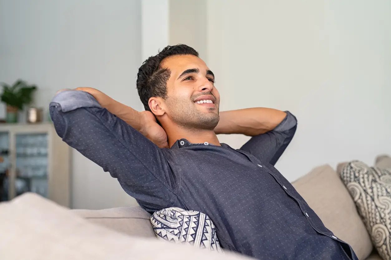 Young indian man at home relaxing and thinking with hands behind head. Middle eastern pensive guy resting at home. Latin thoughtful man sitting on couch while looking up.