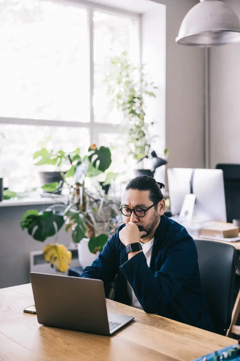 At a creative office workspace, a Japanese art director works on his laptop, with large windows allowing natural light into the office.